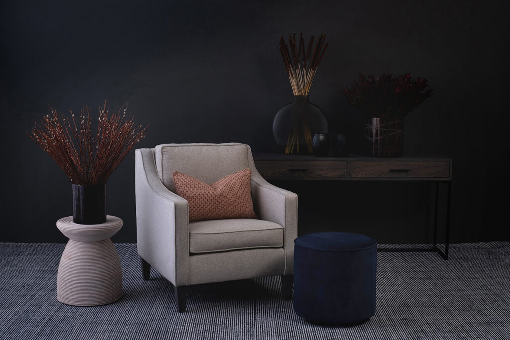 Luxury furniture including a white arm chair on gray rug with wood and metal server behind it, a plush blue ottoman and side table with decor