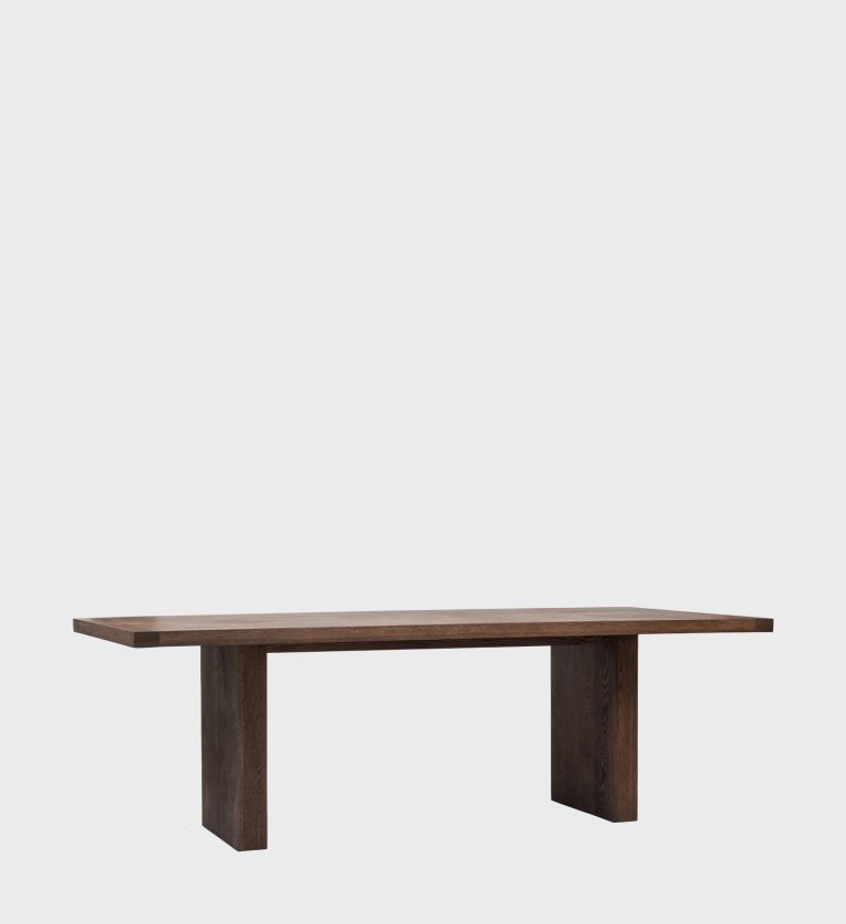 The Sweni Dining Table embodies timeless craftsmanship and understated elegance. Its long rectangular top, made from richly grained dark wood, rests on two solid slab legs that emphasize strength and simplicity.