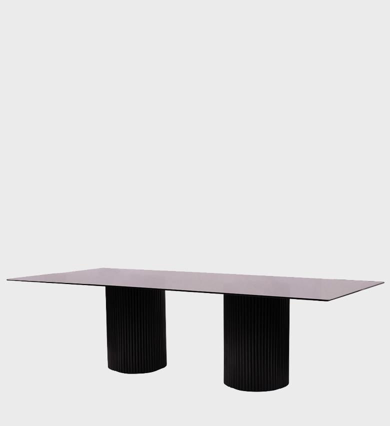 The Fluted Smokey Glass Dining Table is a stunning example of contemporary elegance. Its expansive rectangular top, crafted from smooth smokey glass, rests on two cylindrical fluted bases finished in deep black.