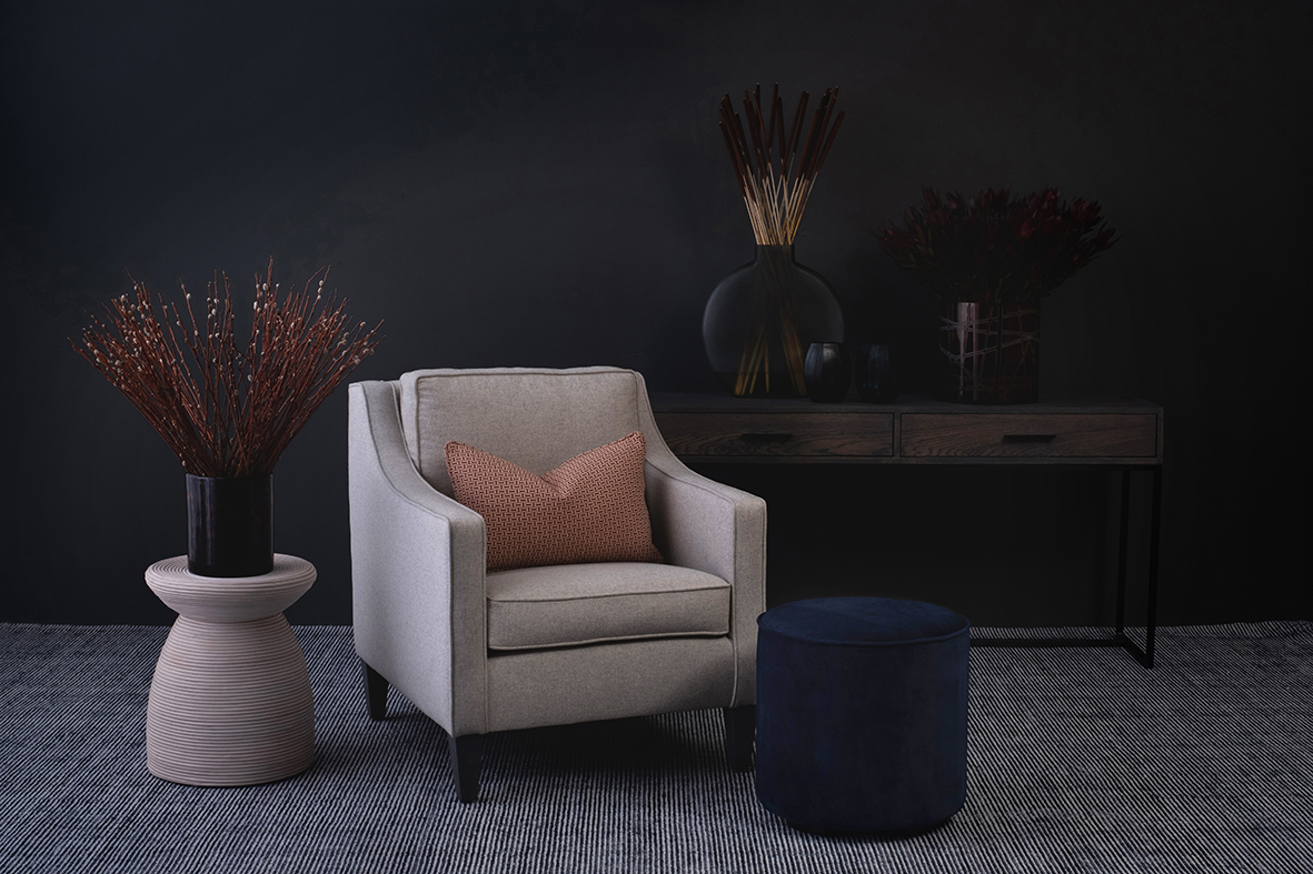 Luxury furniture including a white arm chair on gray rug with wood and metal server behind it, a plush blue ottoman and side table with decor